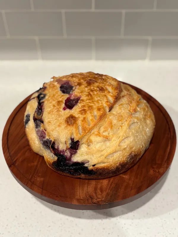 A round, scored loaf with blueberries showing through a golden crust, on a round wooden board