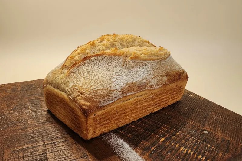 A sourdough loaf baked in a tin, with a flour-dusted split top, sitting on a dark wooden board