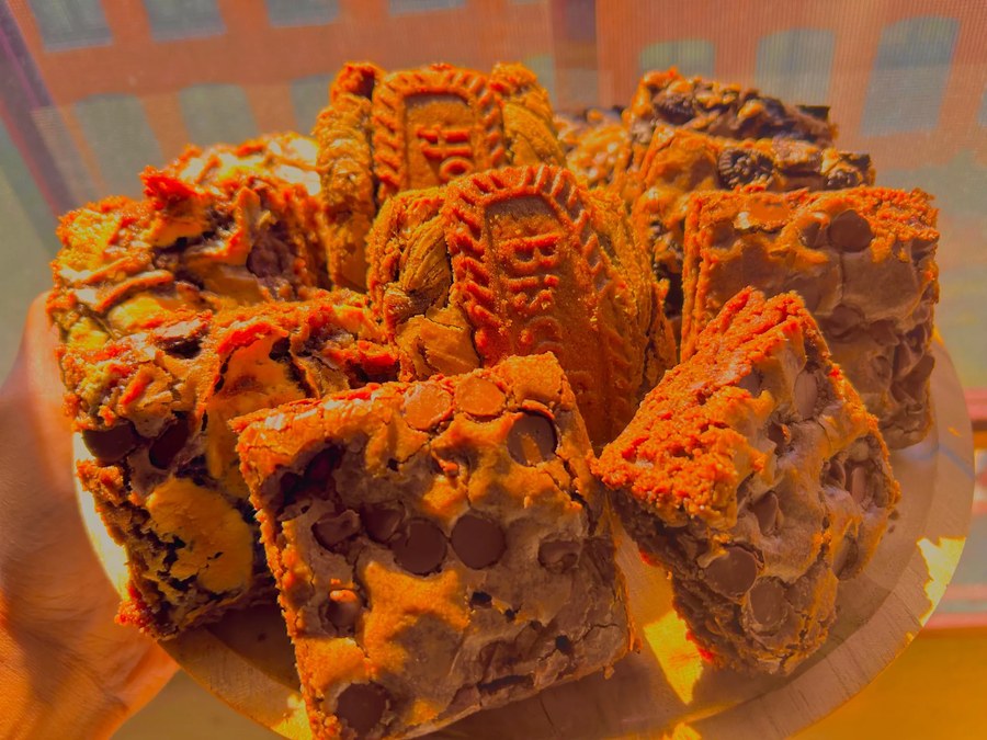 Chocolate chip brownie squares and whole embossed cookies piled on a round wooden board in bright sunlight