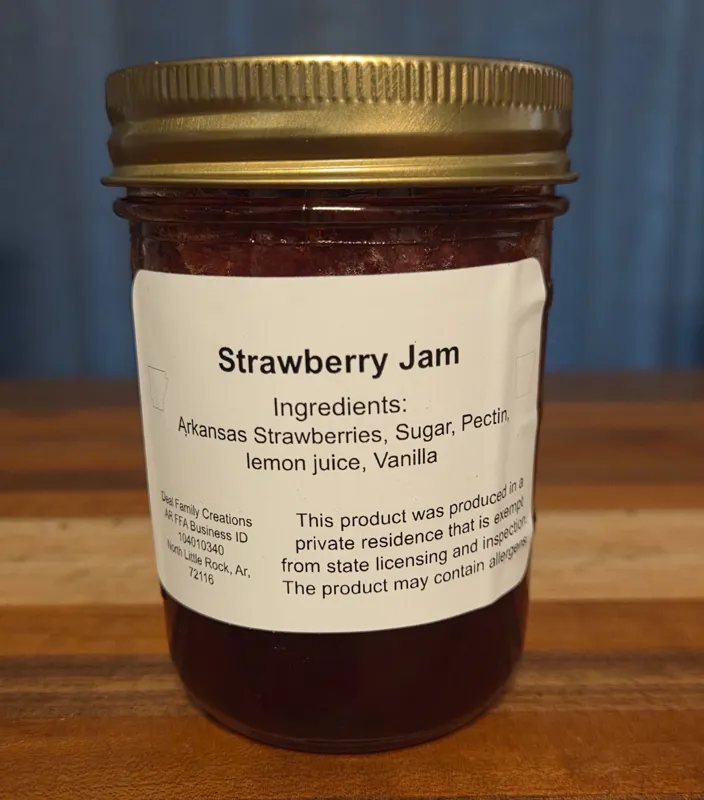 A jar of Strawberry Jam with a white label listing ingredients and a home kitchen statement, on a wood table