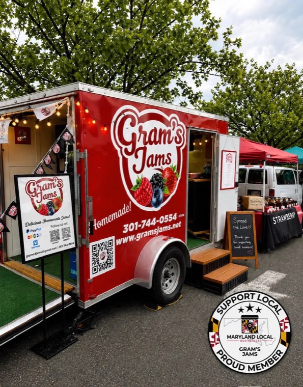A red Gram's Jams trailer parked at an outdoor market, with a sign showing a QR code, a chalkboard sign and a table of jars under a red canopy