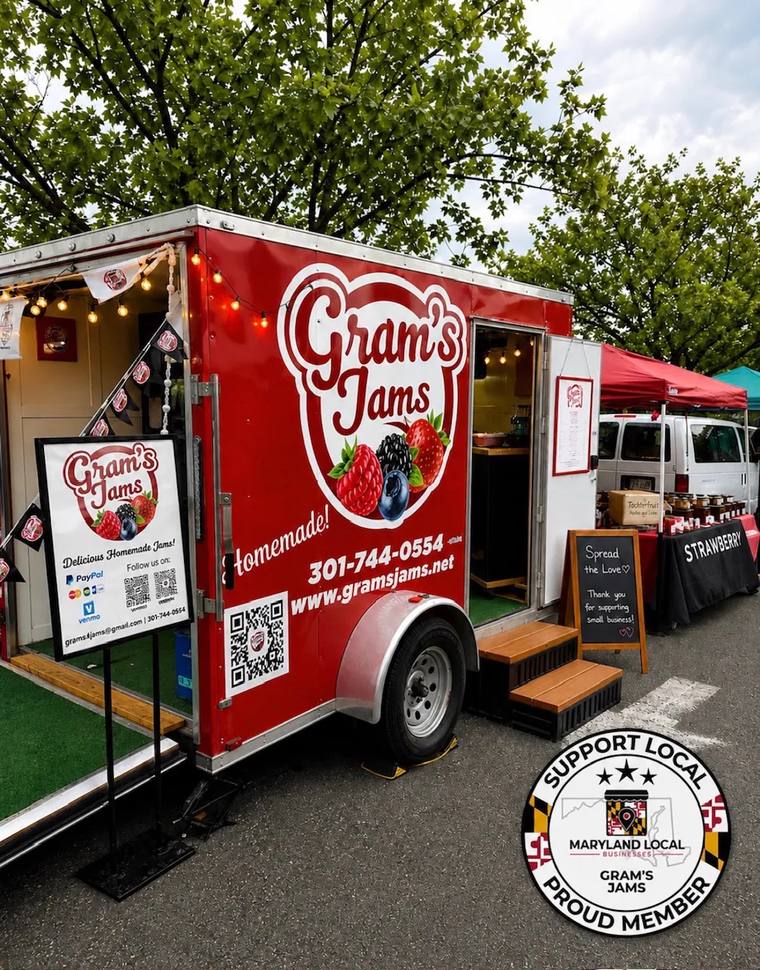 A red Gram's Jams trailer with its side door open, parked beside a red canopy over a table of jars