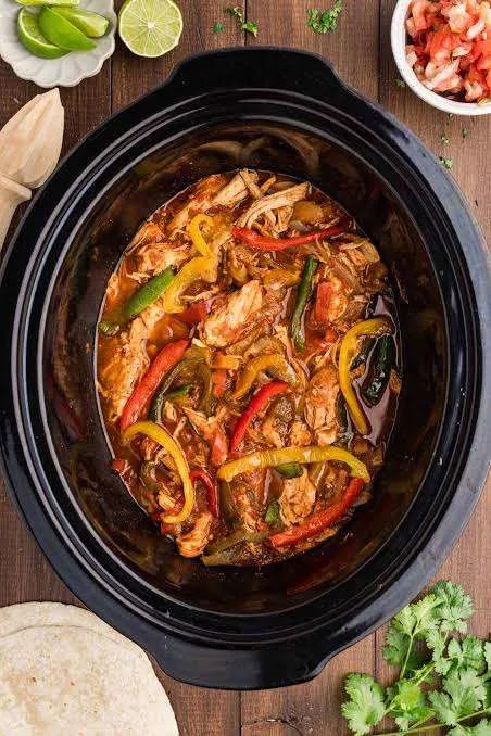 Shredded chicken with sliced red, yellow and green peppers in a black slow cooker, with tortillas, cilantro and lime wedges around it