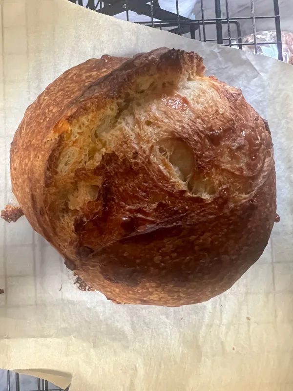 A round sourdough loaf with a dark, glossy, torn crust, cooling on parchment paper over a wire rack