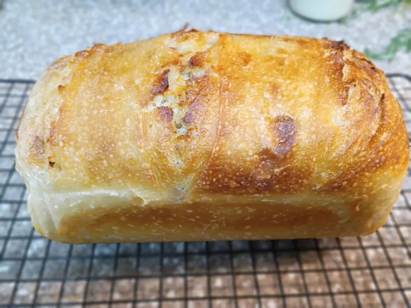 A tall loaf of bread with a golden, scored crust cooling on a wire rack